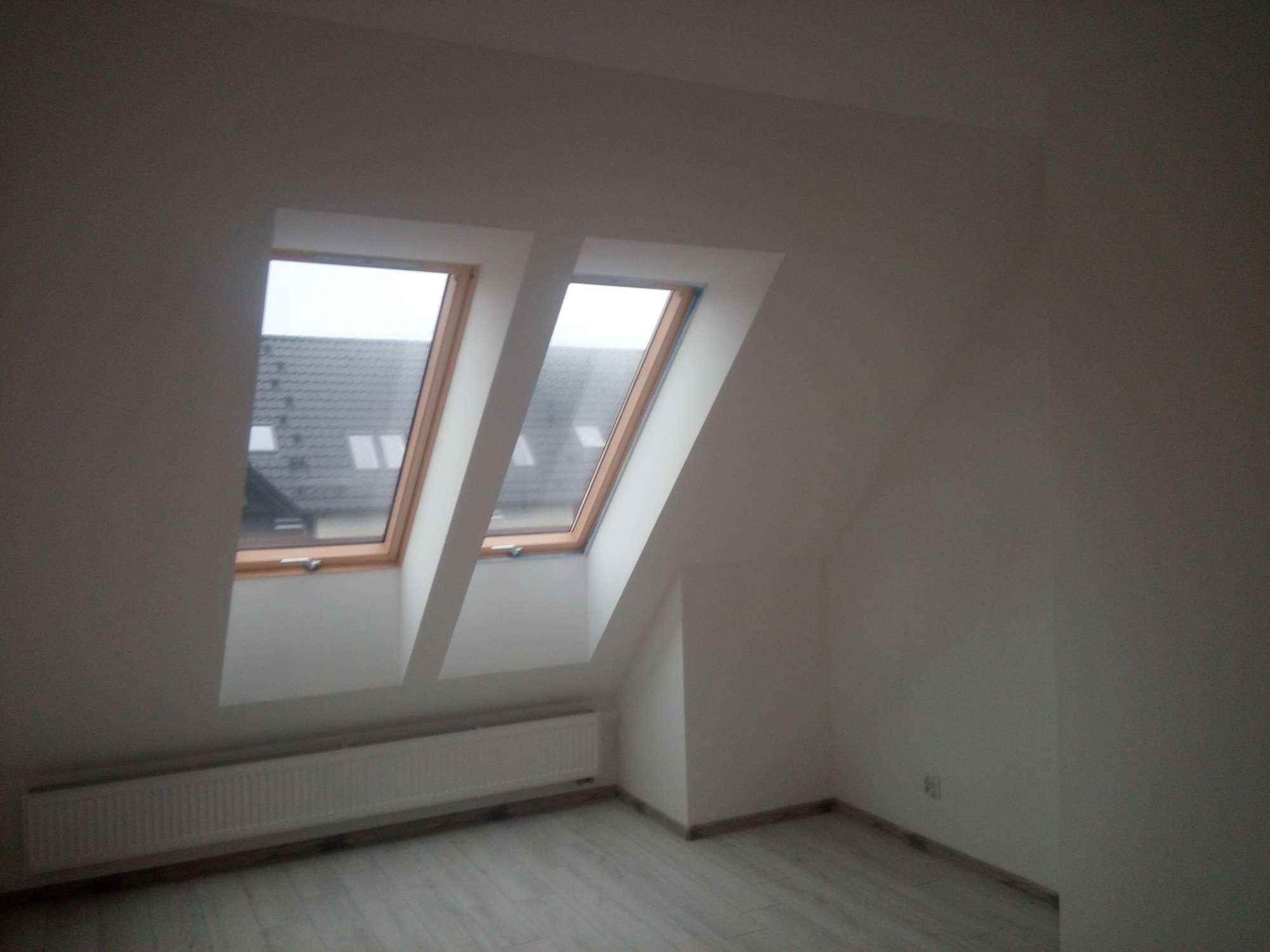 Poddasze z dwoma oknami dachowymi, białe ściany po świeżym wykończeniu, szary panel podłogowy, grzejnik pod oknami.
