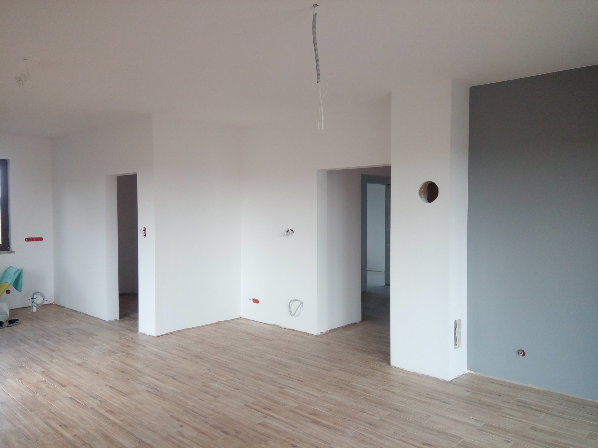 Jasne wnętrze po remoncie z drewnopodobną podłogą, białe ściany częściowo pomalowane na szaro, widoczne puszki elektryczne i wyprowadzone kable, przygotowane do dalszych prac wykończeniowych.