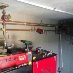 Instalacja grzewcza w kotłowni z widocznym kotłem marki Mocek, miedzianymi rurami, pompami obiegowymi i wiertarką akumulatorową na kotle.