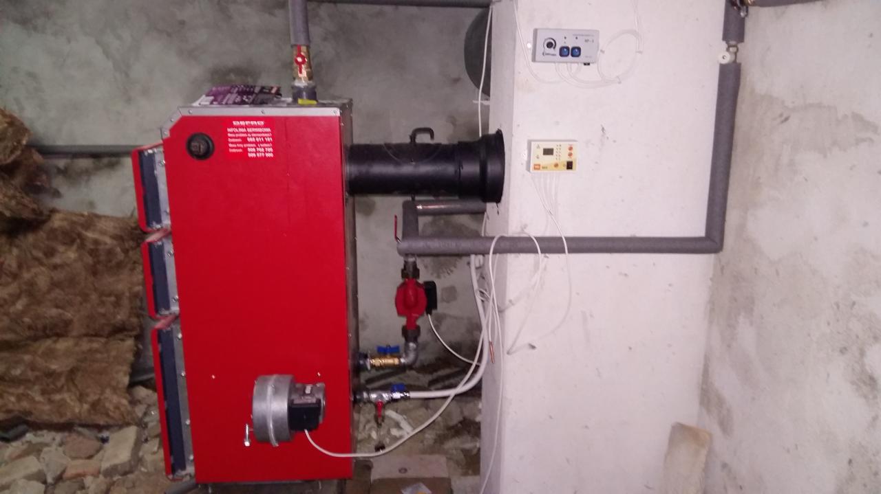 Czerwony piec centralnego ogrzewania z czarnym kominem, widoczne rury instalacji, regulator temperatury na ścianie w surowym pomieszczeniu.