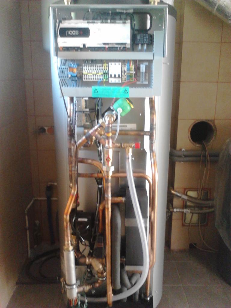 Otwarty panel sterowania nowoczesnego pieca CO z widocznymi przewodami miedzianymi i elementami instalacji w kotłowni wyłożonej kafelkami.