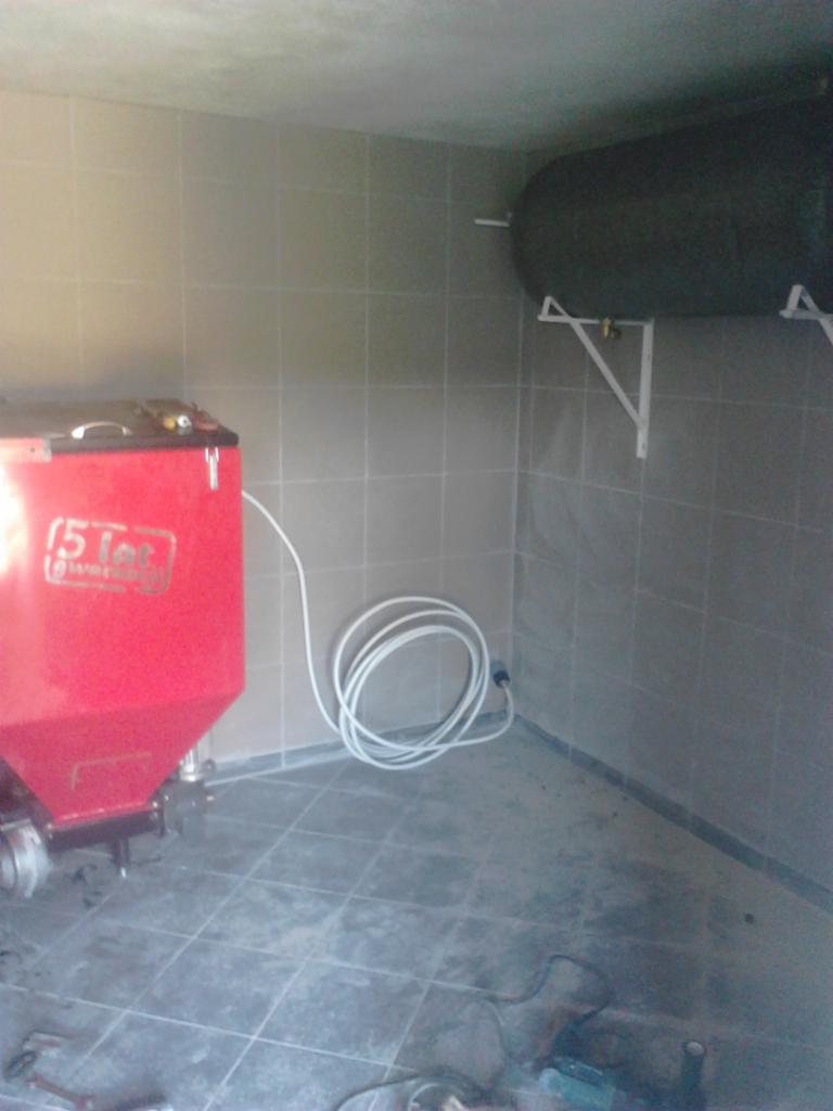 Czerwony piec z podajnikiem na ekogroszek w kotłowni z szarymi płytkami, widoczny zasobnik na wodę na ścianie i zwinięty biały kabel.