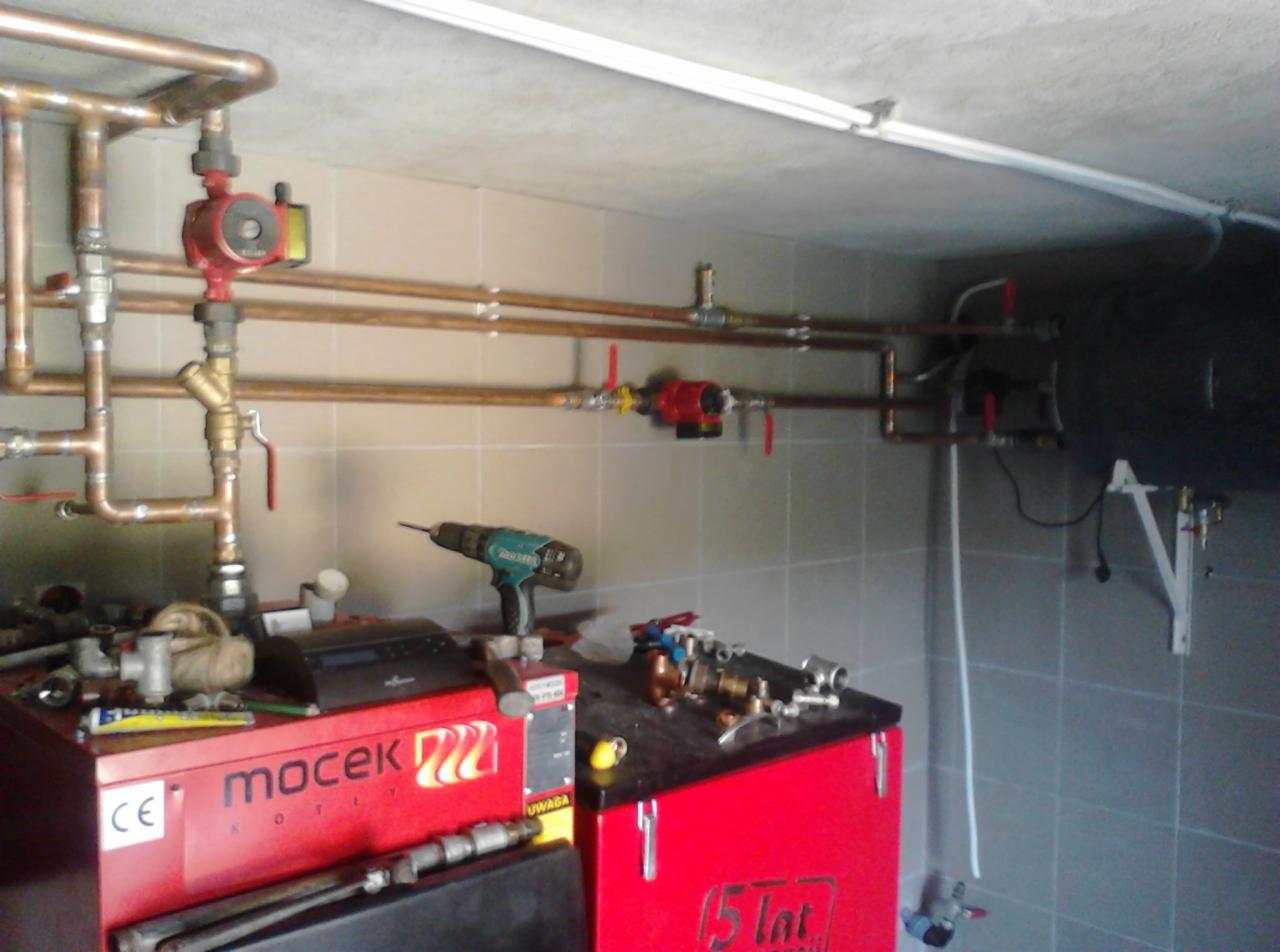 Instalacja grzewcza w kotłowni z widocznym kotłem marki Mocek, miedzianymi rurami, pompami obiegowymi i wiertarką akumulatorową na kotle.