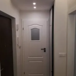 Wnętrze przedpokoju po remoncie: białe ściany, drewnopodobna podłoga, białe drzwi z szybką, sufit podwieszany z oświetleniem punktowym, widoczny fragment szafy przesuwnej.