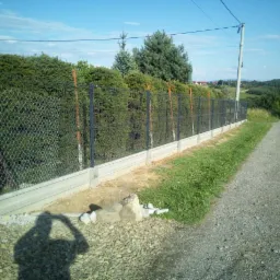 Nowoczesne ogrodzenie z siatki z podmurówką betonową, czarne słupki, widok z boku na tle zieleni i polnej drogi, cień fotografa w lewym dolnym rogu.
