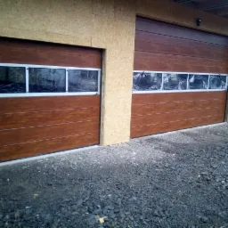 Dwie brązowe segmentowe bramy garażowe z oknami, jeszcze zabezpieczone folią, zamontowane w surowym otworze budynku z płyt OSB, widoczne gruz i rusztowania.