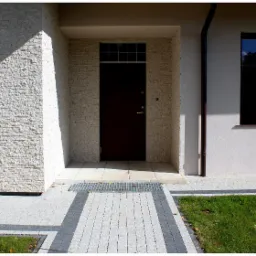 Elewacja budynku z jasnej cegły dekoracyjnej, ciemne drzwi wejściowe z górnym oknem, szara kostka brukowa z pasami ciemniejszej kostki, małe drzewko ozdobne po lewej stronie.