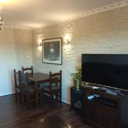 Jasne wnętrze z drewnianym stołem i krzesłami, ściana wykończona płytkami imitującymi kamień, oświetlenie punktowe i dekoracyjny żyrandol.