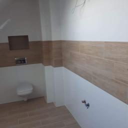 Remonty. Józef. Malowanie - Wykończona łazienka z beżowo-białymi płytkami imitującymi drewno na ścianach i podłodze, wisząca toaleta, wystające przewody elektryczne i rury wodne.