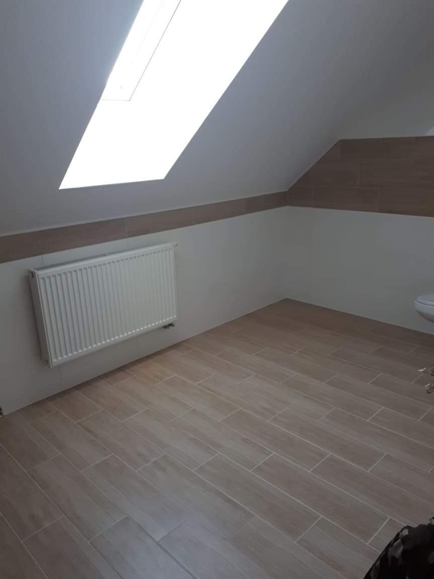 Wykończona łazienka na poddaszu ze skośnym sufitem i oknem dachowym, beżowe płytki podłogowe imitujące drewno, biały grzejnik na ścianie.