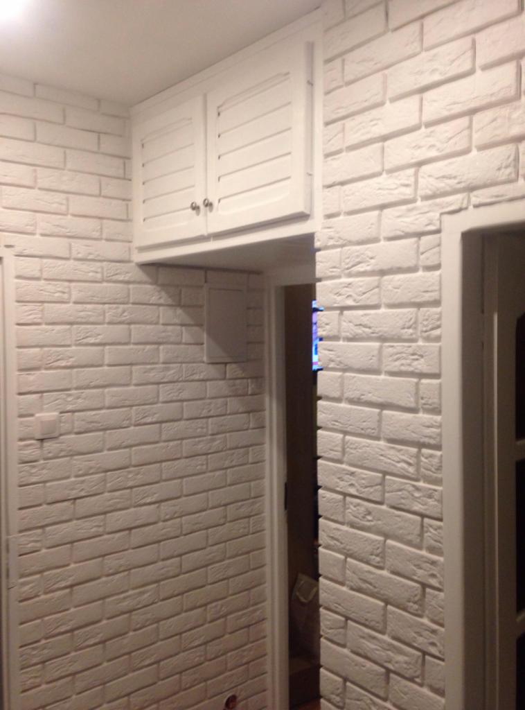 Biała ściana wykończona dekoracyjną cegłą, nad przejściem szafka z białymi drzwiczkami na zawiasach.