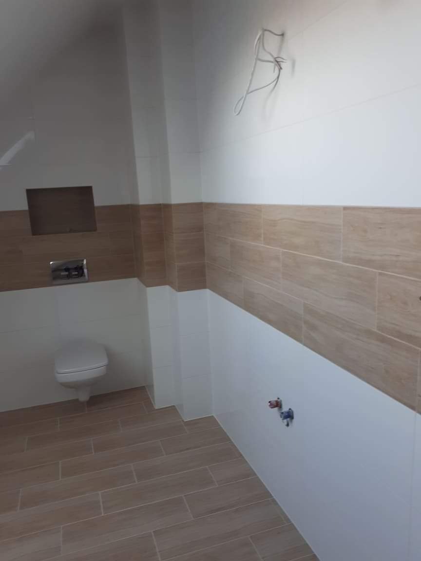 Wykończona łazienka z beżowo-białymi płytkami imitującymi drewno na ścianach i podłodze, wisząca toaleta, wystające przewody elektryczne i rury wodne.