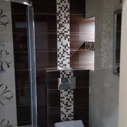 Wykończona łazienka z brązowo-beżowymi płytkami, mozaiką w pionowym pasie, kabiną prysznicową z zaokrąglonym szkłem i podwieszaną toaletą.