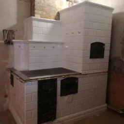 Nowoczesny piec kaflowy z białych płytek, czarne żeliwne drzwiczki i płyta grzewcza, złote detale, surowe wykończenie ściany w tle.