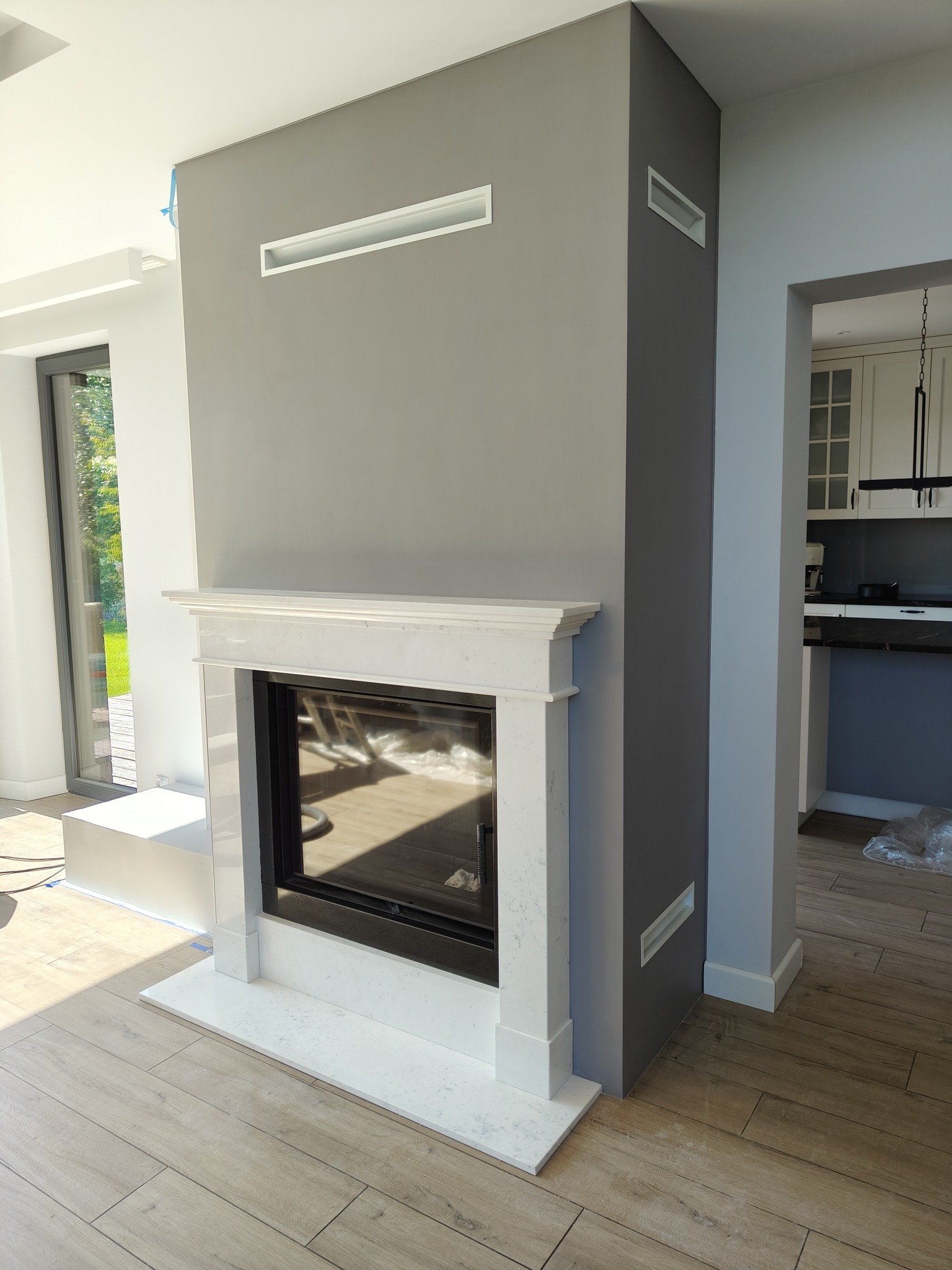 Nowoczesny kominek z białym marmurem i czarnym wkładem, wkomponowany w szarą ścianę z minimalistycznymi kratkami wentylacyjnymi, na tle jasnego wnętrza.