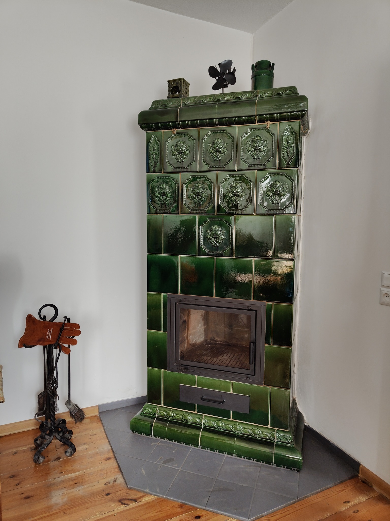 Zielony piec kaflowy z ozdobnymi kaflami i czarnymi drzwiczkami, stojący w rogu pomieszczenia z drewnianą podłogą i akcesoriami kominkowymi obok.