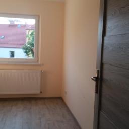 Mirosław Staśkiewicz - Pomalowane na brzoskwiniowo ściany pokoju z szarą podłogą imitującą drewno, biały grzejnik pod oknem i ciemne drzwi z klamką po prawej stronie.