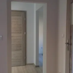 Wnętrze mieszkania po remoncie: widok na korytarz z dwiema drewnopodobnymi drzwiami i jasną podłogą, ściany pomalowane na szaro, oświetlenie punktowe w suficie.