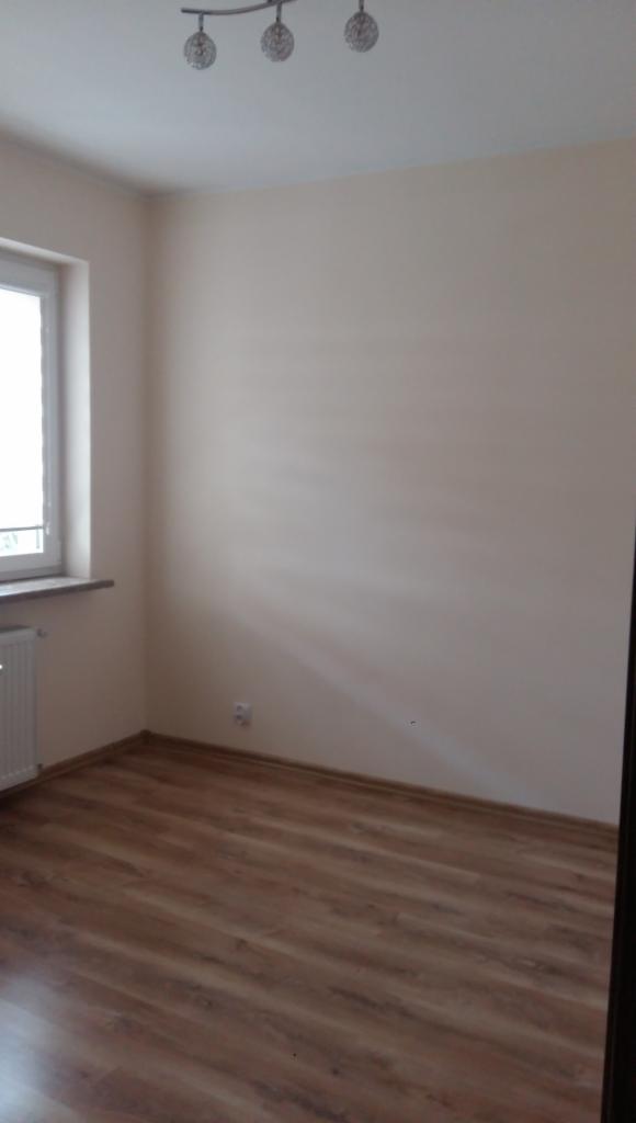 Pusty pokój z panelami podłogowymi w odcieniach brązu, kremowe ściany, okno z białymi ramami i grzejnik oraz potrójna lampa sufitowa z metalowymi kloszami.