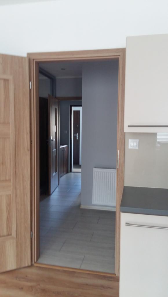 Wnętrze mieszkania po remoncie: widok z kuchni na korytarz z szarą ścianą, grzejnikiem i otwartymi drzwiami do kolejnych pomieszczeń, podłoga wyłożona panelami i płytkami imitującymi drewno.