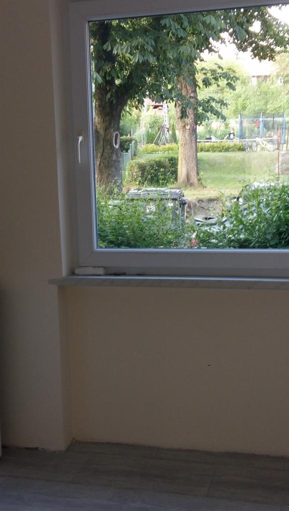 Świeżo pomalowana ściana w jasnym odcieniu, widok na ogród przez okno z białymi ramami, zarys podłogi z szarych płytek.