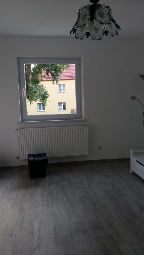 Pusty pokój z szarą podłogą imitującą drewno, białymi ścianami, grzejnikiem pod oknem z widokiem na budynek z czerwonym dachem i drzewo, oraz ozdobną lampą sufitową.