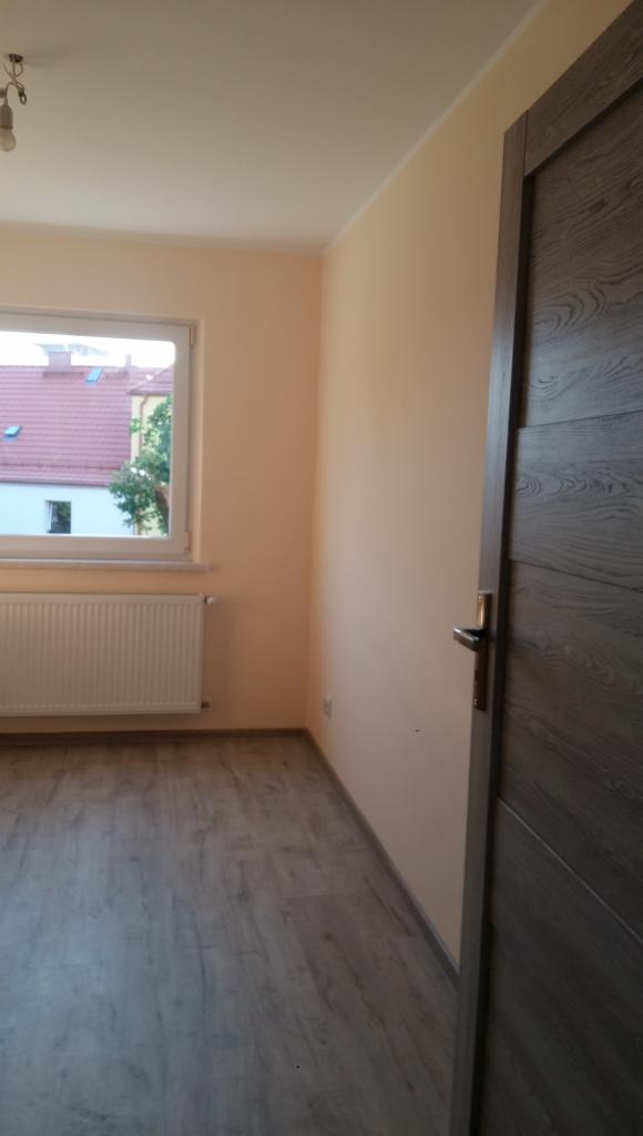 Pomalowane na brzoskwiniowo ściany pokoju z szarą podłogą imitującą drewno, biały grzejnik pod oknem i ciemne drzwi z klamką po prawej stronie.