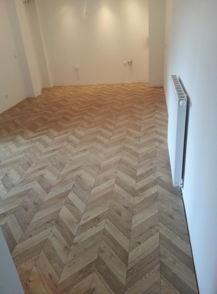 Nowo ułożona podłoga z paneli w jodełkę, widoczny biały grzejnik na ścianie i puste, białe ściany.