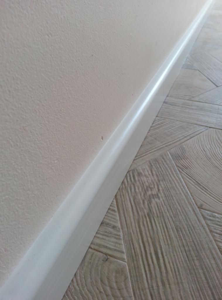 Biała listwa przypodłogowa zamontowana przy jasnej ścianie i podłodze z paneli ułożonych w jodełkę.