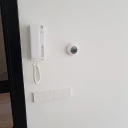 Biała ściana z zamontowanym domofonem, okrągłym termostatem i poczwórnym włącznikiem światła