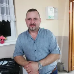 Mężczyzna w niebieskiej koszuli opiera się o głośnik, w tle okno z firanką i apteczka pierwszej pomocy. Uśmiechnięty, z zarostem i zegarkiem na ręku.