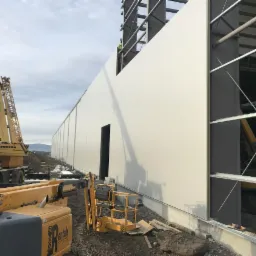 Budowa hali z widoczną ścianą z płyt warstwowych, żurawiem Liebherr i podnośnikiem koszowym Rentals Group na placu budowy.