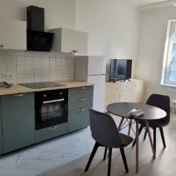 Nowoczesna kuchnia z zielonymi szafkami, drewnianym blatem i marmurową podłogą w jasnym mieszkaniu z widokiem na budynek mieszkalny.