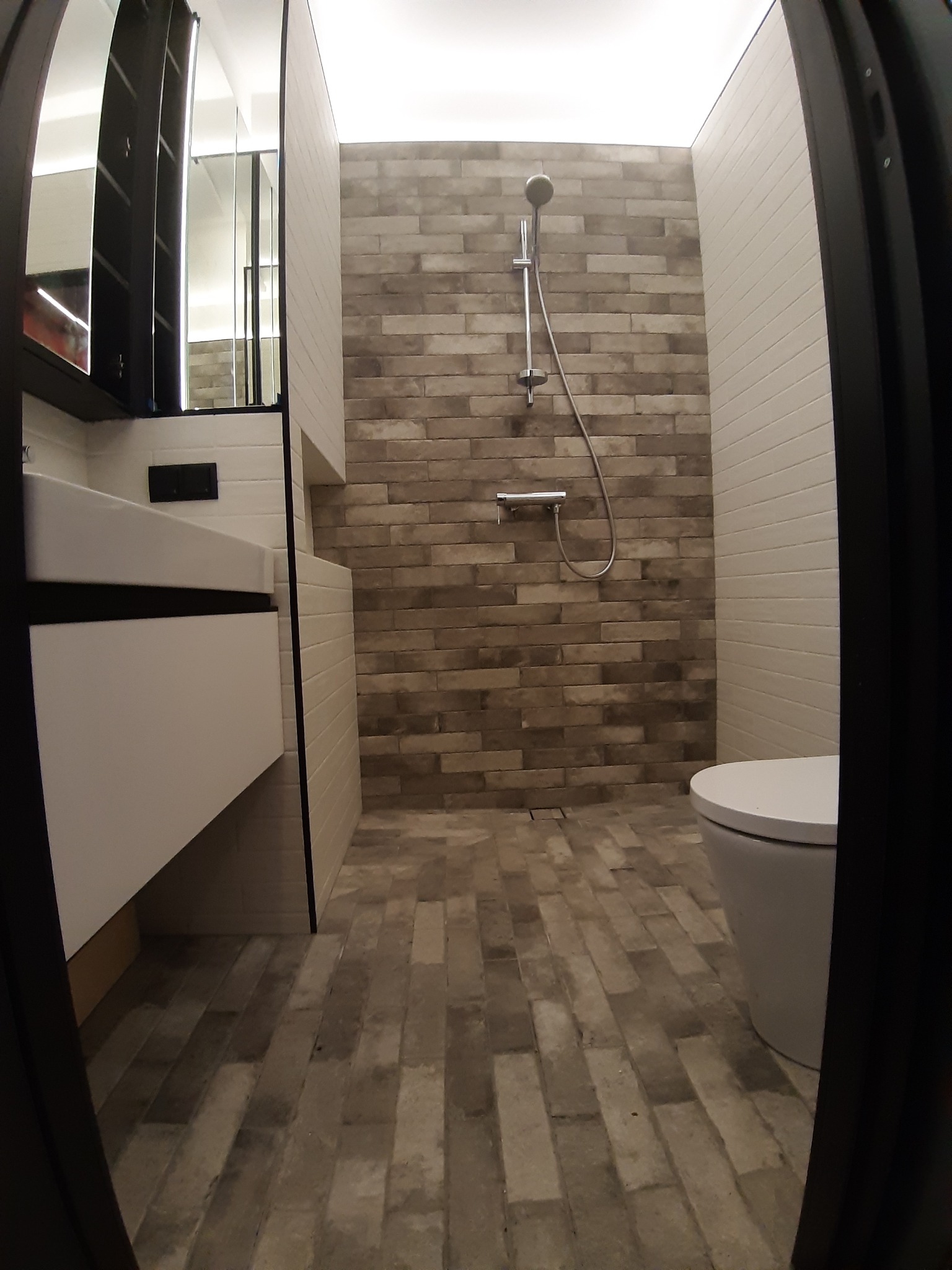 Nowoczesna łazienka z kabiną prysznicową typu walk-in, wykończona płytkami imitującymi cegłę w odcieniach szarości i beżu, z białą umywalką nablatową i toaletą, widok zza futryny drzwi.