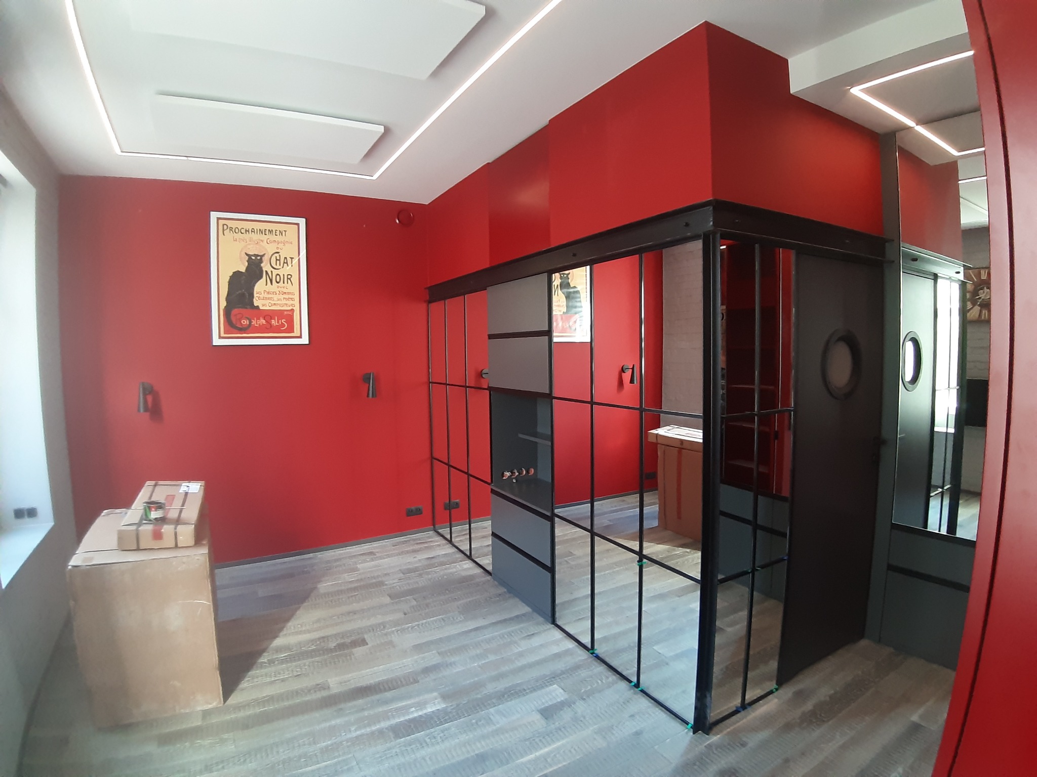 Pomieszczenie z czerwonymi ścianami, drewnianą podłogą i czarną, przeszkloną konstrukcją oddzielającą przestrzeń. Na ścianie plakat vintage z czarnym kotem. Na podłodze kartony z materiałami...