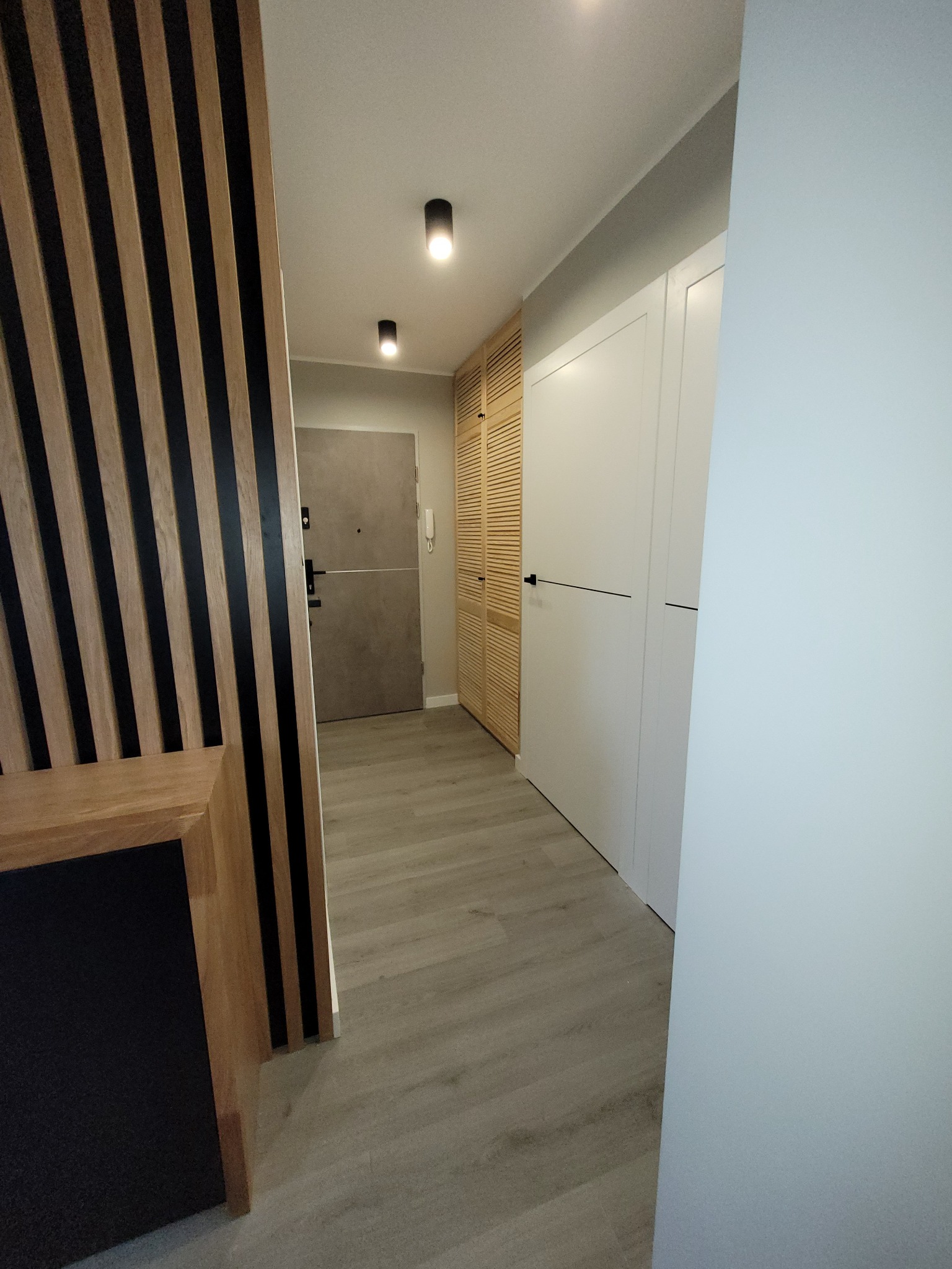 Nowoczesny przedpokój z drewnianymi lamelami na ścianie, szarą podłogą imitującą drewno, białymi szafami w zabudowie i szarymi drzwiami wejściowymi.