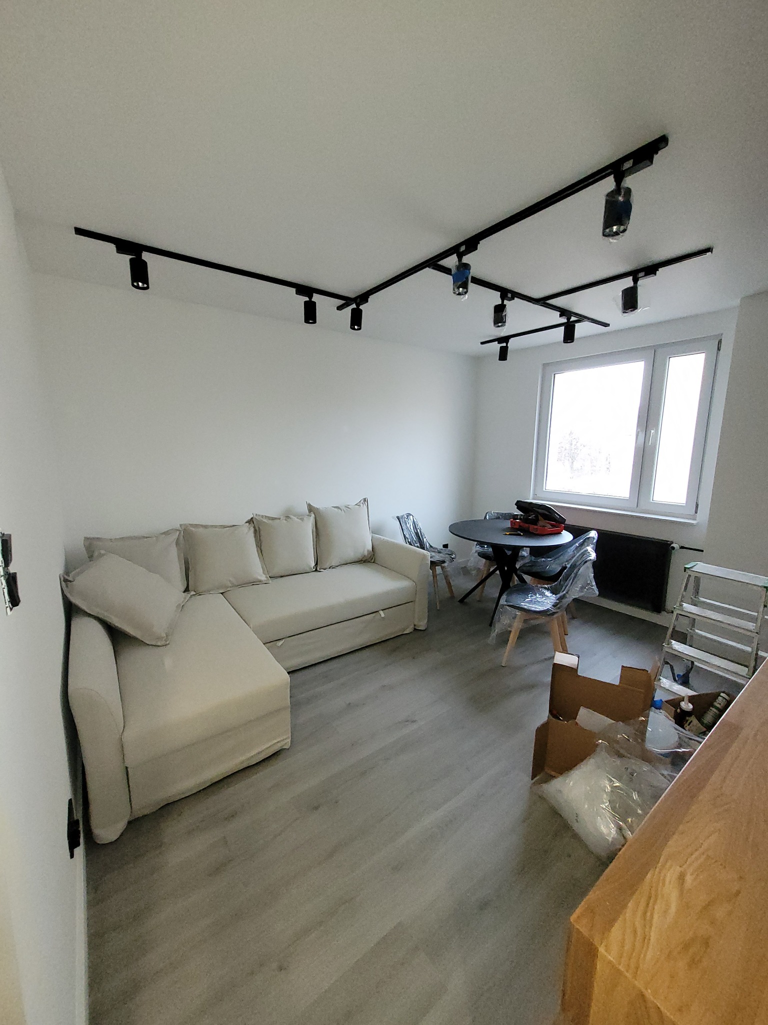 Jasny salon z narożną sofą, okrągłym stołem i nowoczesnym systemem oświetlenia szynowego na suficie. Krzesła przykryte folią ochronną, drabina oparta o ścianę, kartony na podłodze.