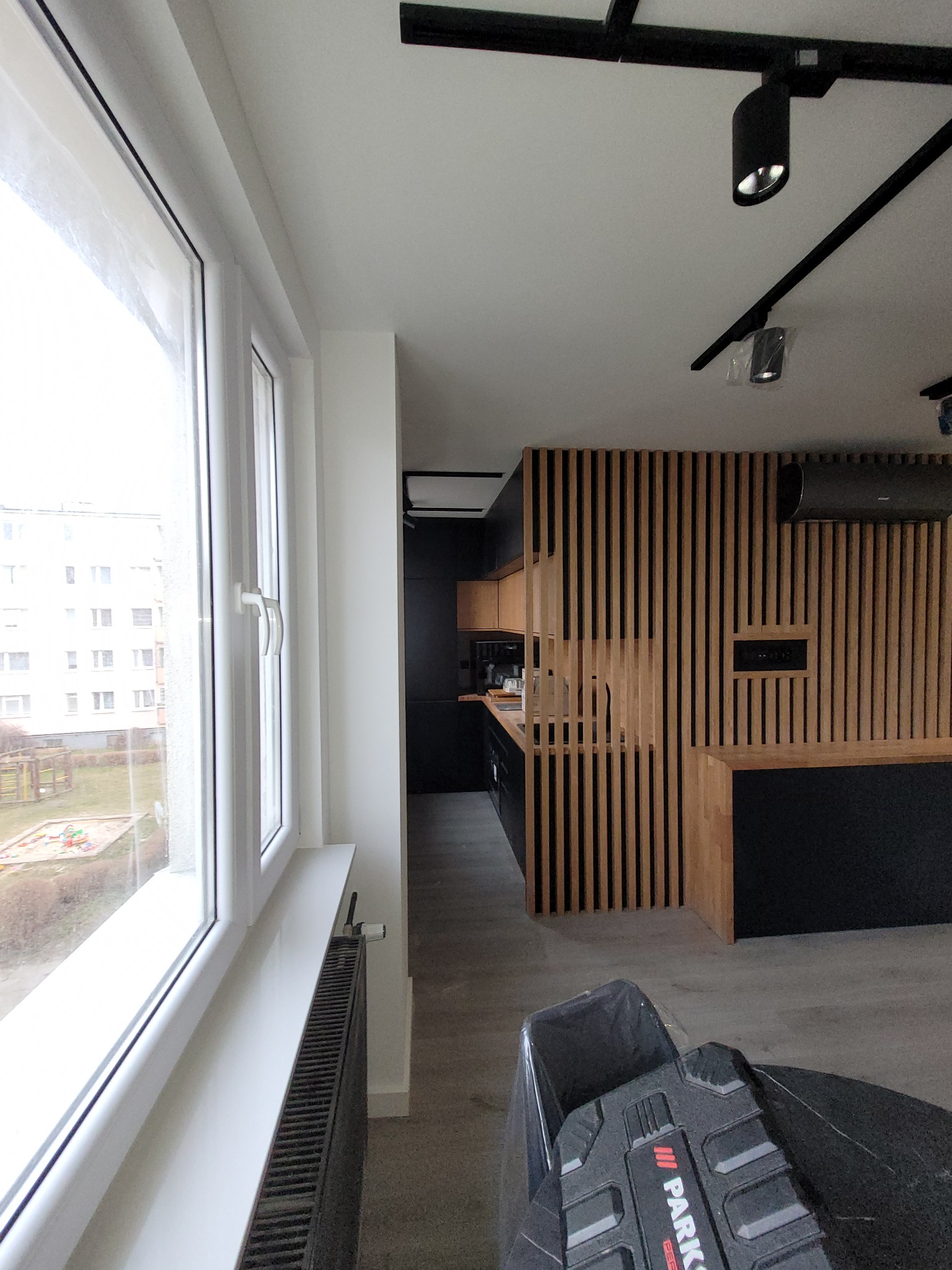 Nowoczesna kuchnia z drewnianą ścianką działową z pionowych listew, widok zza okna z białymi ramami i grzejnikiem, na pierwszym planie fragment odkurzacza.