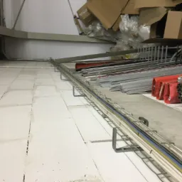 Metalowa korytka kablowa z ułożonymi przewodami elektrycznymi w różnych kolorach, zamocowana na ścianie w surowym wnętrzu, z widocznymi materiałami budowlanymi i kartonami w tle.