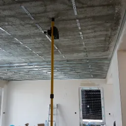 Montaż stelaża sufitu podwieszanego z płyt kartonowo-gipsowych, widoczny laser krzyżowy na żółtym statywie, surowy betonowy strop z zamocowanymi elementami konstrukcji.