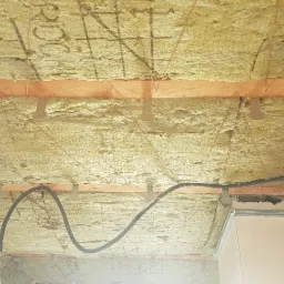 Poddasze ocieplone wełną mineralną, widoczne drewniane belki, metalowe uchwyty mocujące, kable w peszlu i fragment ściany.