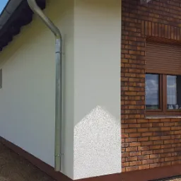 Pracownik zabezpiecza elewację budynku z białą strukturą tynku i ceglaną wstawką wokół okna, widoczny fragment rynny i zaparkowany samochód dostawczy z narzędziami.