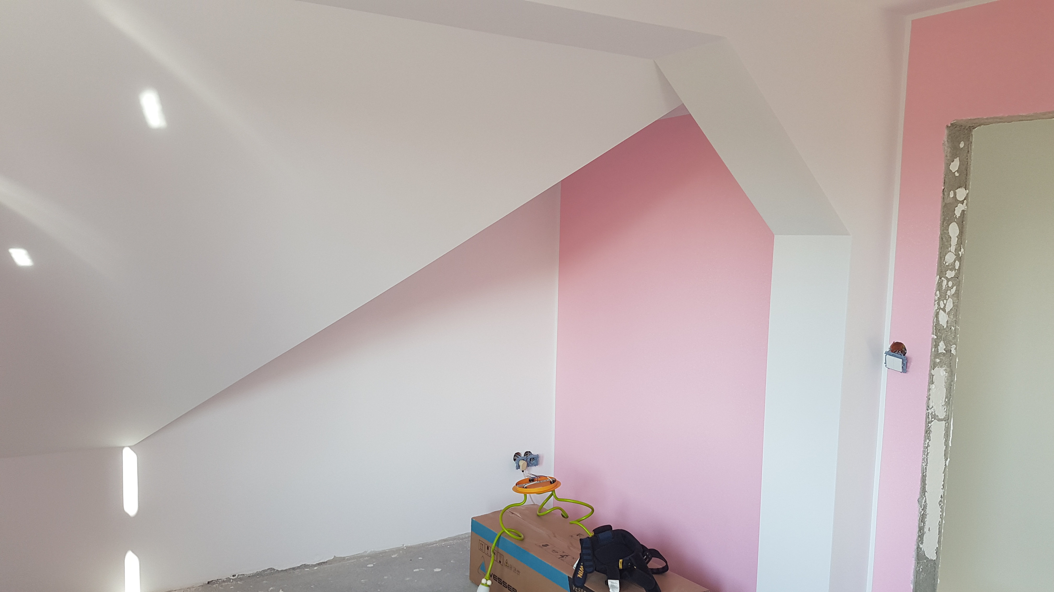 Poddasze po malowaniu: białe skosy i fragment różowej ściany, na podłodze karton z narzędziami i przedłużaczem, widoczne gniazdko elektryczne.