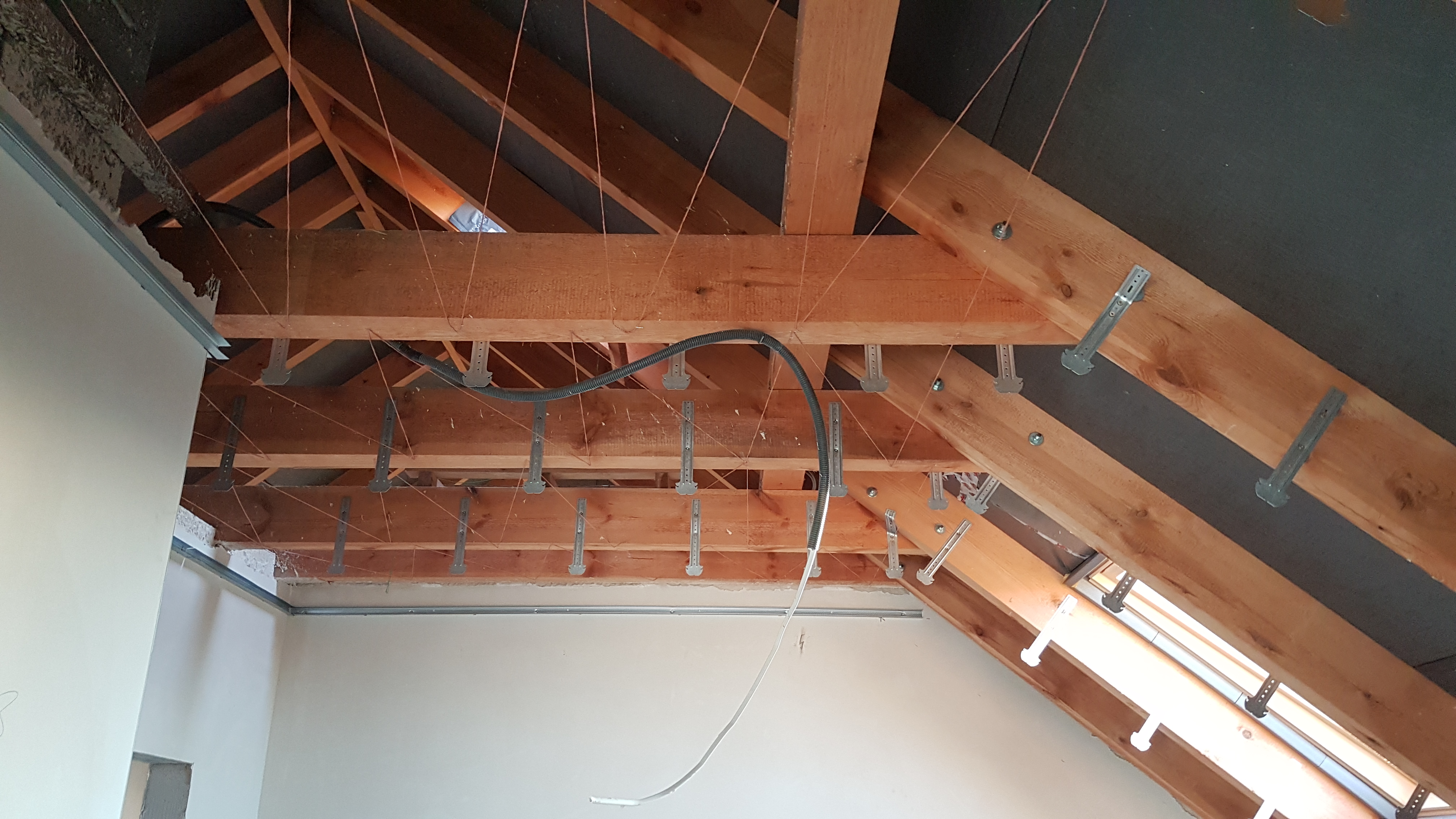Konstrukcja drewniana poddasza z zamocowanymi metalowymi wieszakami, widoczne kable i fragment ściany w trakcie prac wykończeniowych.