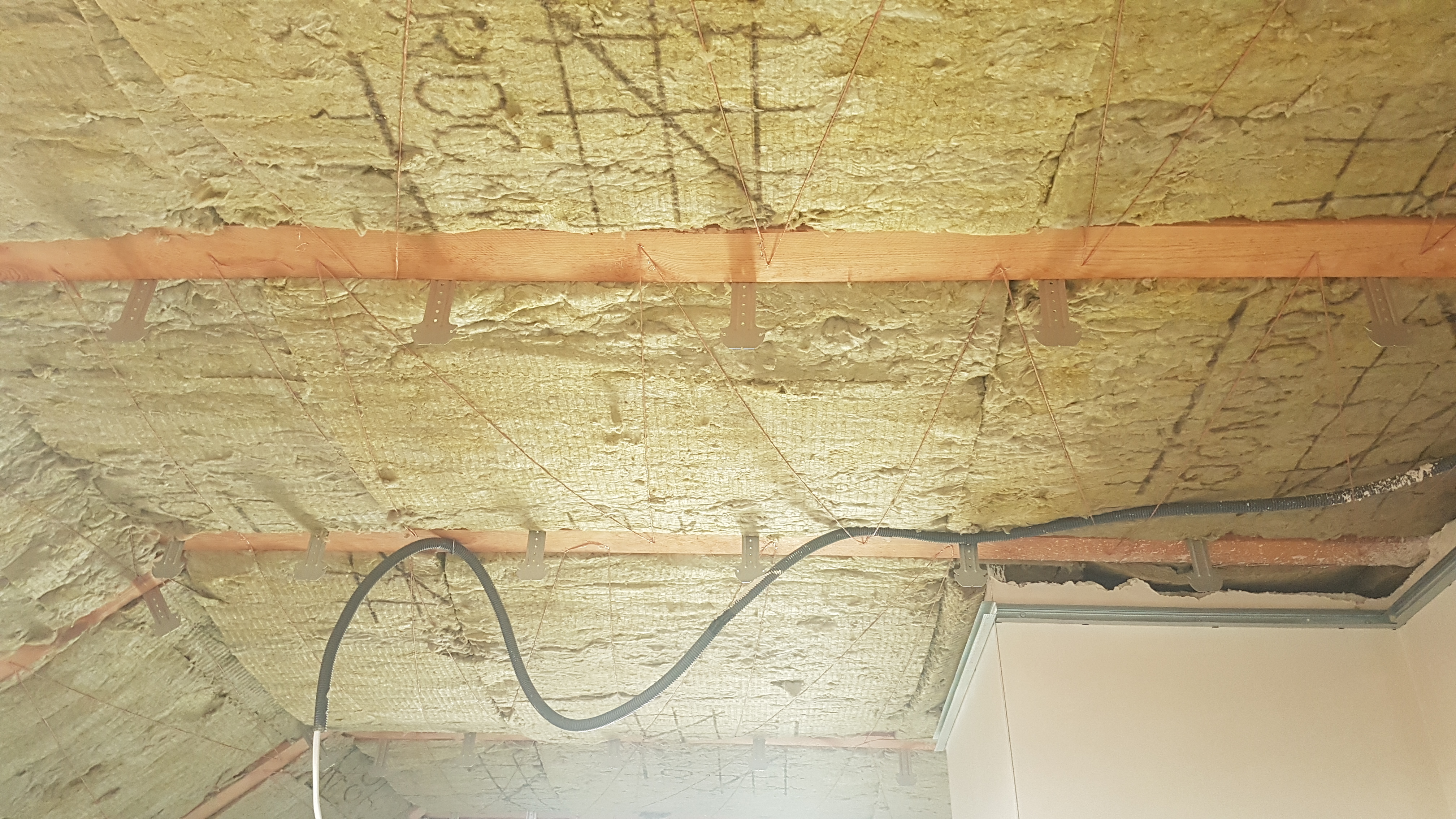 Poddasze ocieplone wełną mineralną, widoczne drewniane belki, metalowe uchwyty mocujące, kable w peszlu i fragment ściany.