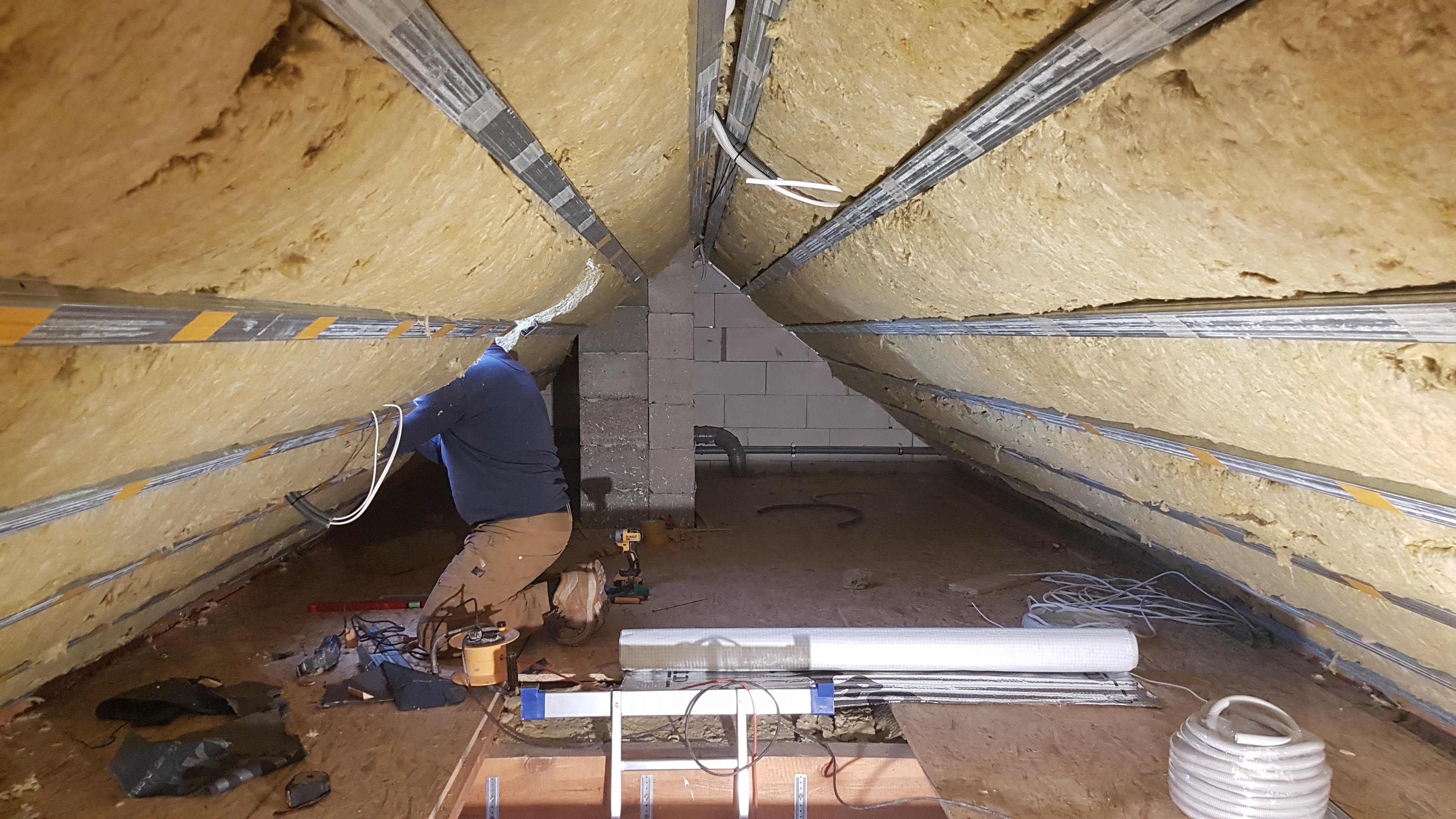 Pracownik na kolanach montuje instalację elektryczną w ocieplonym wełną mineralną poddaszu z widocznymi profilami metalowymi i narzędziami pracy.