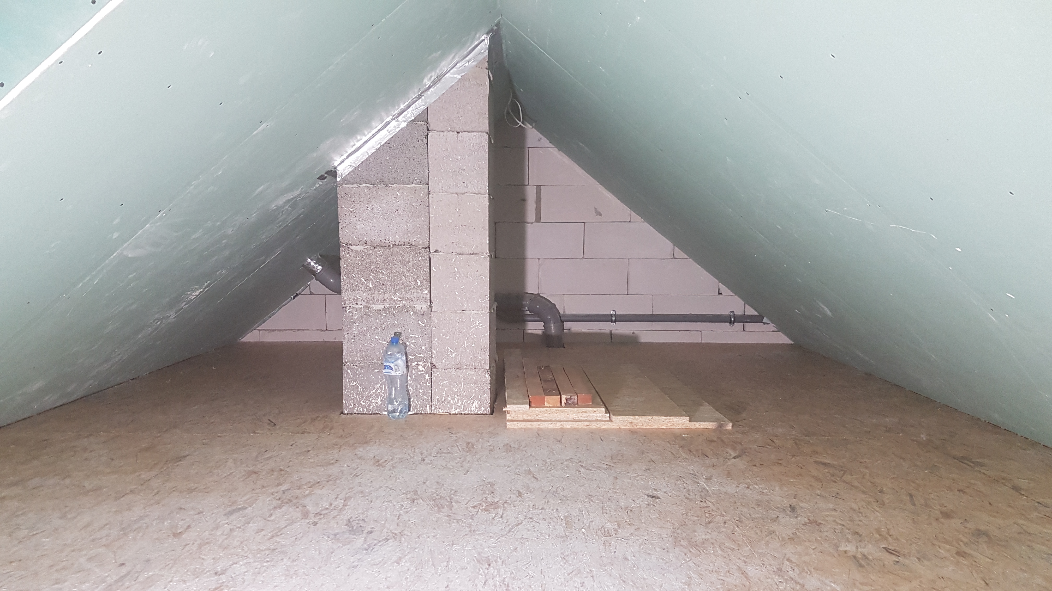 Poddasze w trakcie adaptacji: płyty karton-gips zamontowane na skosach, fragment komina z pustaków, butelka wody na podłodze z płyt OSB, widoczne przewody instalacyjne.