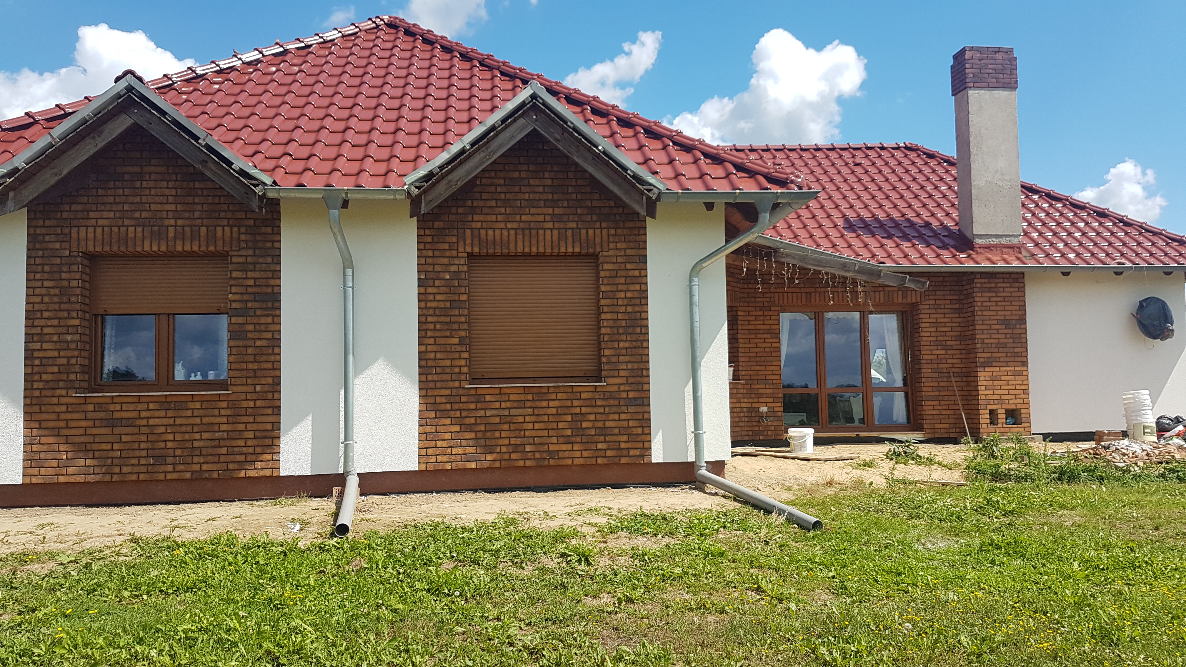Bryła domu jednorodzinnego z czerwoną dachówką, fragment elewacji z cegły klinkierowej, okna z brązowymi roletami zewnętrznymi i szarymi rynnami spustowymi, trawnik z przodu budynku.