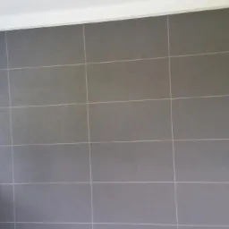 Ściana wyłożona szarymi, matowymi płytkami ceramicznymi o regularnym kształcie i jasnych fugach, widok z bliska.