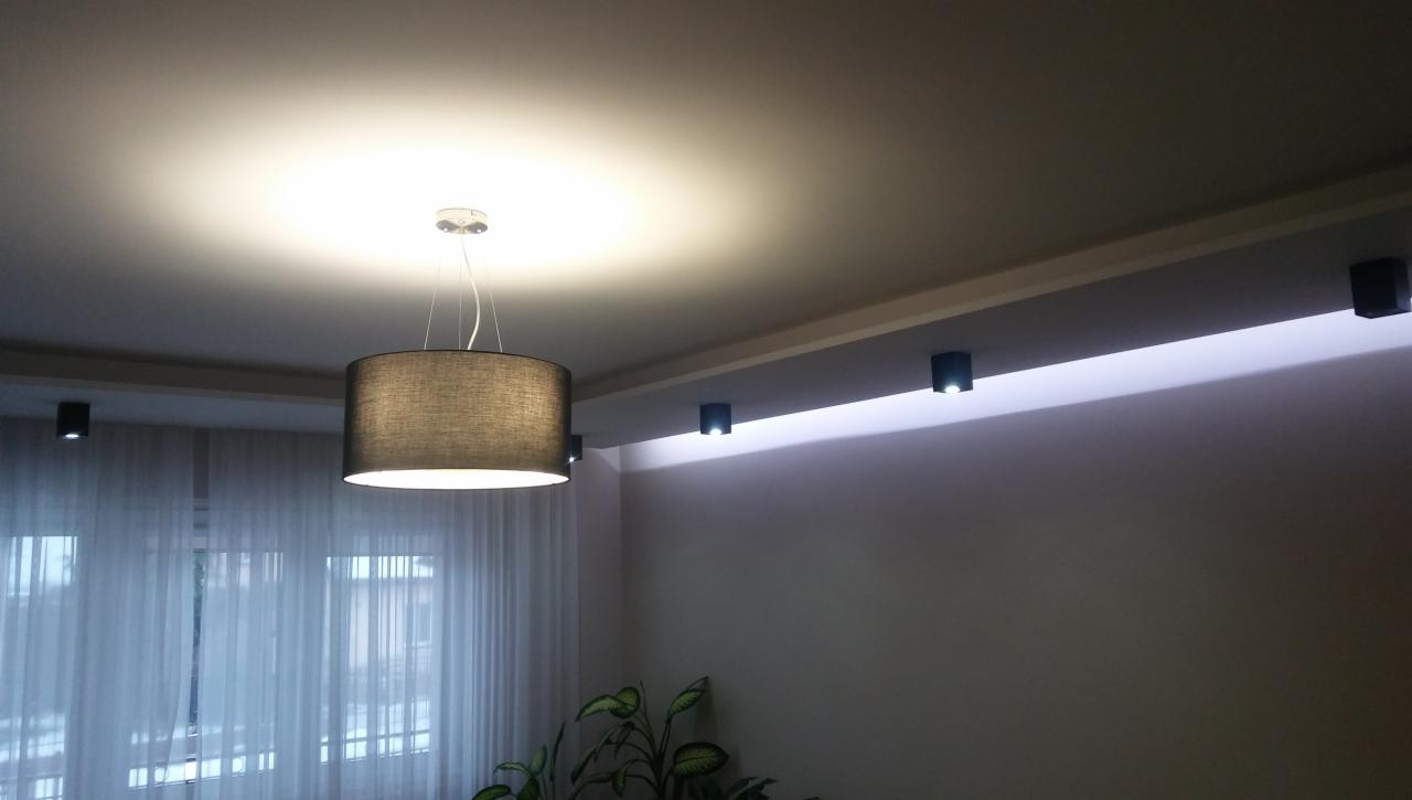 Nowoczesne oświetlenie sufitowe: wisząca lampa z abażurem i listwa z punktowymi reflektorami LED, subtelne światło w minimalistycznym wnętrzu.